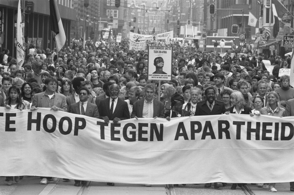 Black and white photograph of protestors in a flag-lined street, holding a banner that partially reads 'hoop tegen apartheid'.