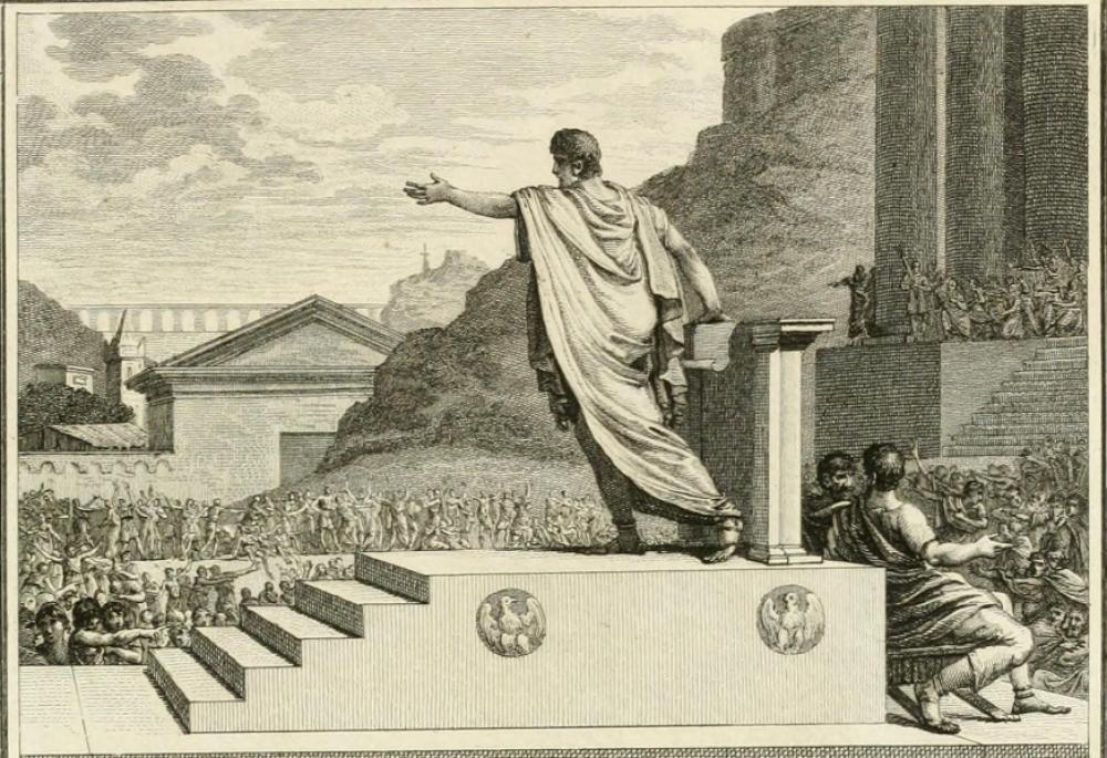 A man wears a Roman style toga and sandals standing at a podium, standing atop a a platform with five steps and two circular eagle emblems. Hundreds of people, similarly dressed, surround the speaker