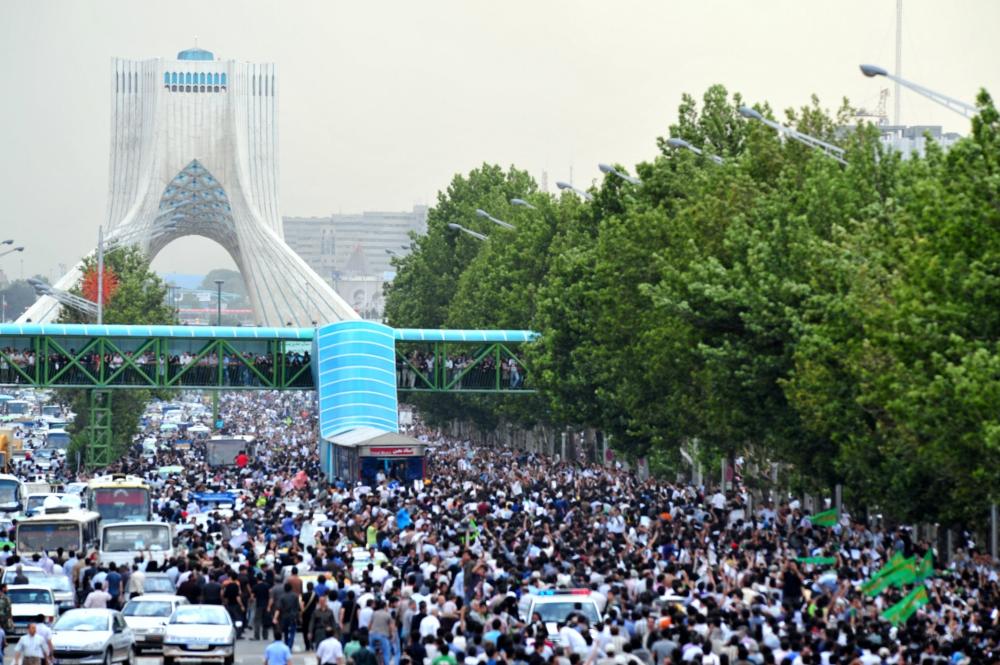 Cityscape filled with people on streets, set of trees lining the road and a large bridge in the background. 