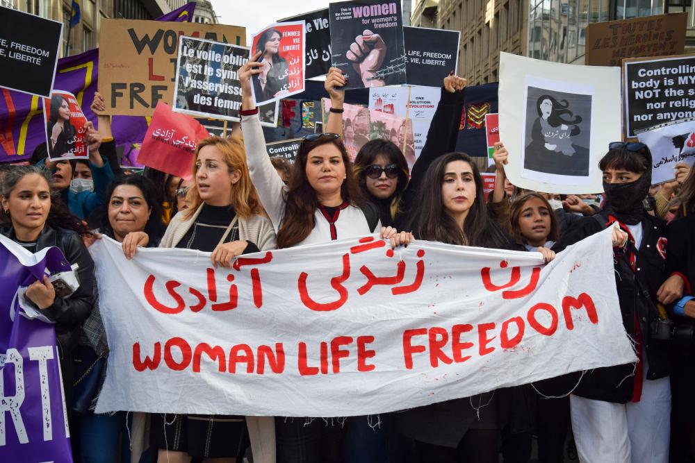 Photo of young female protestors holding signage, a white banner with red lettering that says "Woman Life Freedom" 