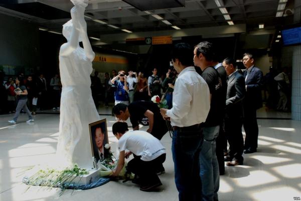 Onlookers holding flowers and cameras gather near a white female-figured statue, two men kneel to lay flowers at statue's feet and photo of regal man